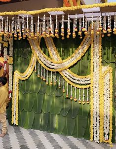 two men standing in front of a wall decorated with yellow and white flowers, garlands and beads