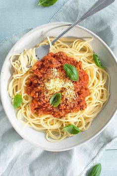 a white bowl filled with spaghetti and sauce on top of a blue table cloth next to a spoon