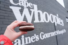 a hand holding a chocolate covered doughnut in front of a sign that says cape whoops