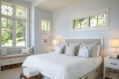 a white bed sitting in a bedroom next to two windows with lots of pillows on top of it