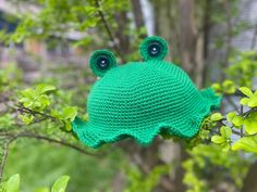 a crocheted frog hat hanging on a tree branch with green leaves in the background