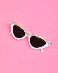 a pair of white sunglasses with crystals on the top and bottom, against a pink background