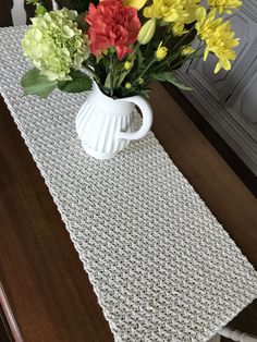 a crocheted table runner with flowers in a vase on it, sitting on a dining room table