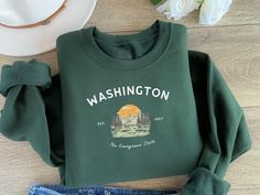 This high quality, sustainably made Washington State crewneck is the coziest gift to remember all your adventurous memories of the Evergreen State. Featuring the classic phrase "WASHINGTON est 1967 The Evergreen State," this crewneck is perfect for anyone who loves Washington's natural beauty and laid-back vibes. Crafted from 80% ring spun cotton and 20% polyester, the Gildan® SF000 sweatshirt offers a super soft feel with its brushed interior and 100% ring spun cotton face. The medium-heavy fabric (8.4 oz/yd²) makes it perfect for chilly days or cozy nights. With its dropped shoulder design for a relaxed fit, this crewneck will quickly become your go-to. We've made sure that style and comfort align with eco-conscious production. The sweatshirt is made with OEKO-TEX certified low-impact dy State Crewneck, Adventurous Travel, Evergreen State, Hiker Gifts, Cozy Gift, Gifts For Nature Lovers, Shoulder Design, Washington State, Travel Gifts