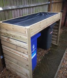 a wooden structure with a blue trash can in the middle and a black plastic bin under it