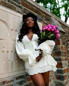 a woman in a white dress and hat leaning against a brick wall with purple flowers