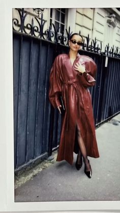 a woman standing on the sidewalk in front of a fence wearing a long leather coat