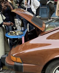 people standing around a brown car with its hood open and food on the table in front of it