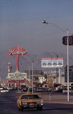 cars driving down the road in front of buildings and billboards with mountains in the background