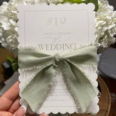 a person holding up a wedding card with a bow on it and flowers in the background