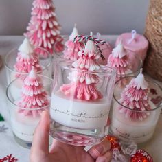 a person holding a glass with some pink candles in it and small christmas trees on the side