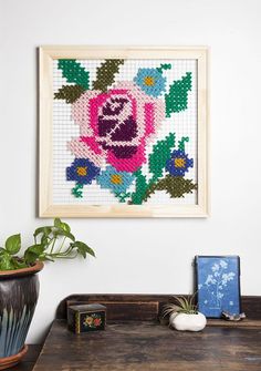 a cross stitch picture hanging on the wall next to a potted plant