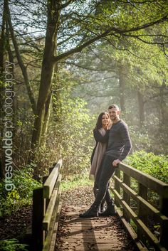 a man and woman standing on a bridge in the woods with their arms around each other