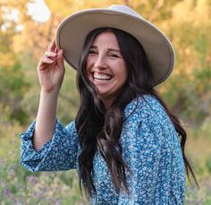 a woman wearing a hat smiles while standing in a field with wildflowers behind her