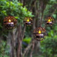 three hanging lanterns with lit candles in them are suspended from chains and surrounded by trees