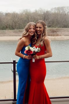 two beautiful women standing next to each other on a pier near the water with flowers in their hands