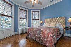 a bedroom with blue walls and wooden floors has a bed, two windows, and a ceiling fan