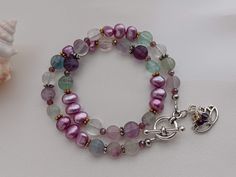 This is a lovely wrap bracelet I made with semi-precious freshwater pearls in a satin grape color (dyed) and Flourite faceted coin stones.  Flourite is an interesting stone that comes in a wide range of colors but most commonly in clear, green, and purple. It is believed to cleanse and purify the body and stabilize energy.   This bracelet consists of a long beaded strand that will wrap twice around your wrist.  The semi-precious beads range from 7 to 8mm in size.  Metal components are made of pewter except for the Lotus flower charm which is made of a zinc-based alloy.  The closure is a pewter round toggle clasp. A pretty lotus flower charm and 2 wire-wrapped crystals dangle from the closure as well. This bracelet will fit wrists measuring up to 7 inches.    This is a handmade piece. All m Gift Single Strand Pearl Bracelet With Round Beads, Double Strand Gemstone Beads Bracelet As Gift, Spiritual Single Strand Beaded Bracelets As Gift, Spiritual Single Strand Beaded Bracelet As Gift, Gemstone Beads Wrap Bracelet As Gift, Diy Necklace Patterns, Wrapped Crystals, Bracelet Pearls, Grape Color