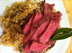 a white plate topped with meat and rice on top of a wooden table next to a green leaf