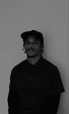 a black and white photo of a man with a mustache wearing a hat, standing against a wall