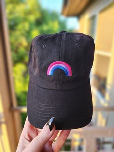 a person holding up a black hat with a rainbow embroidered on the front and side