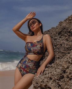 a woman in a bathing suit leaning against a rock on the beach with her hands behind her head