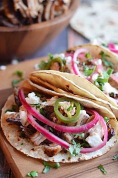 two tacos with meat, onions and peppers on a cutting board next to a bowl