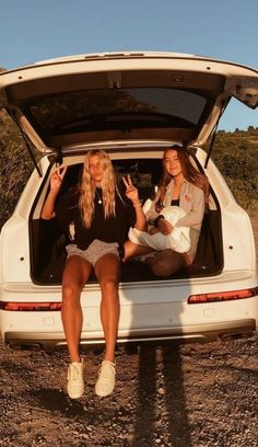 two women sitting in the back of a car with their trunk open and one holding her hand up