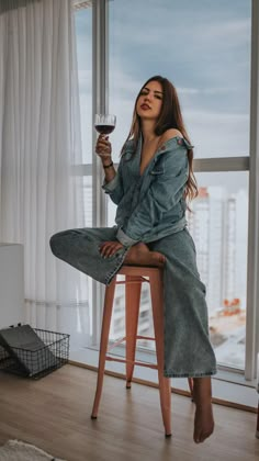 a woman sitting on a stool holding a glass of wine in front of a window