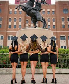 four girls in black graduation gowns are standing near a horse statue with their backs turned to the camera