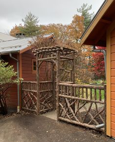 a wooden gate made out of branches in front of a brown building with trees around it