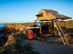 a truck parked next to the ocean with a tent on it's roof and ladder