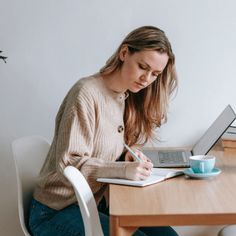 a woman sitting at a wooden table writing on a notepad with a laptop in front of her