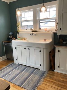 a white kitchen sink sitting under two windows next to a rug on top of a wooden floor