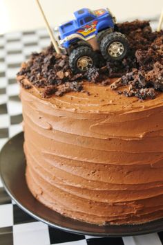 a birthday cake with chocolate frosting and monster trucks on top, sitting on a checkered tablecloth