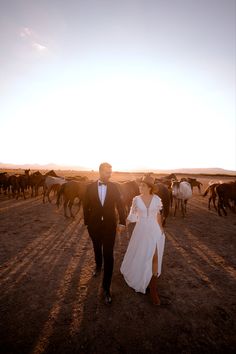 a man and woman are walking in the middle of a field with horses behind them