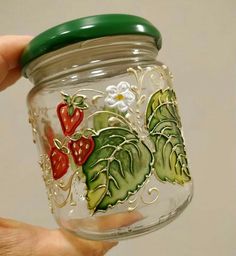 a hand holding a glass jar with strawberries and leaves painted on the side,