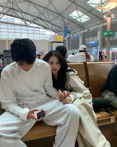 two people sitting next to each other on a bench in an airport terminal, one holding a cell phone