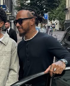 two men standing next to each other in front of a car on a city street