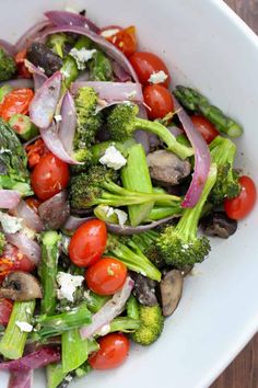 a salad with broccoli, tomatoes, onions and mushrooms in a white bowl