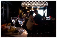 a man and woman standing next to each other in front of a bar with wine glasses