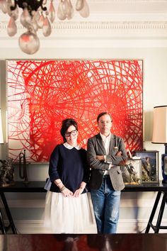 two people standing next to each other in front of a large red piece of art