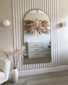 a white chair sitting in front of a mirror next to a vase filled with dry grass