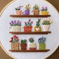 a cross stitched wall hanging with potted plants and flowers on wooden shelfs