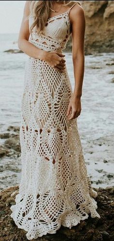 a woman standing on top of a rock next to the ocean wearing a white crocheted dress