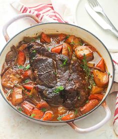 a pot filled with beef and carrots on top of a white table cloth next to silverware