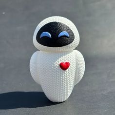 a white knitted toy with a black face and red heart on its chest sitting on a gray surface