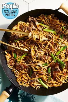 a wok filled with noodles and meat on top of a blue table cloth next to chopsticks