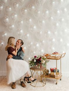 two people sitting on a chair in front of a wall with white balloons and flowers