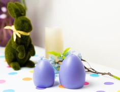 two purple eggs sitting on top of a table next to a teddy bear and candle